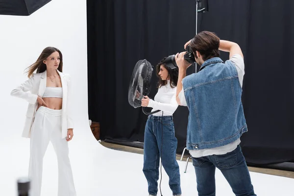 Модель в белом костюме позирует рядом с фотографом и ассистентом с электрическим вентилятором — стоковое фото