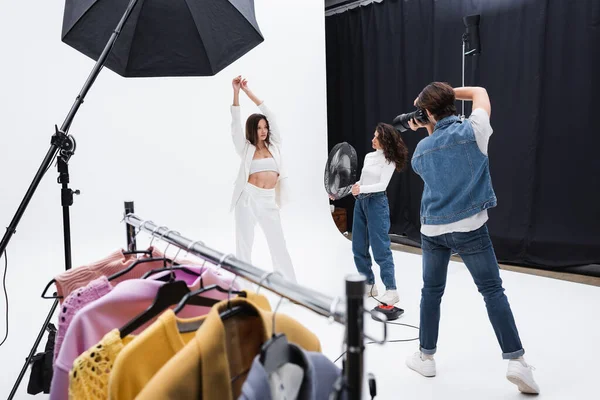 Trendy model posing with raised hands near photographer, assistant with electric fan and rack with clothes — Stock Photo