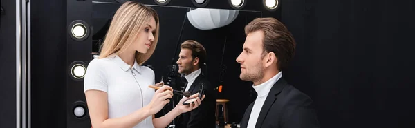 Maquilladora rubia sosteniendo cepillo cosmético y polvo facial cerca del hombre en la sala de maquillaje, pancarta - foto de stock