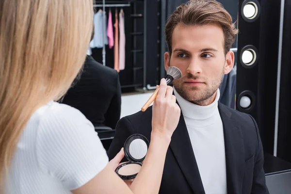 Artista de rostro borroso aplicando polvo facial con cepillo cosmético en el hombre en el estudio de fotos - foto de stock