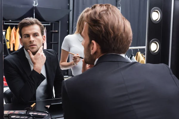 Joven tocando la cara mientras se mira en el espejo cerca de artista de maquillaje - foto de stock