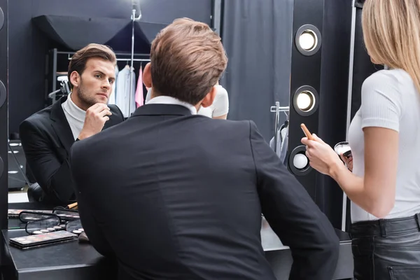 Visage artista sosteniendo la cara en polvo cerca del hombre mirando en el espejo en la sala de maquillaje - foto de stock