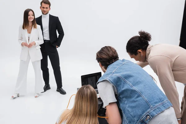 Lächelnde Models beim Anblick des multiethnischen Teams, das in der Nähe des Laptops im Fotostudio auf Weiß arbeitet — Stockfoto