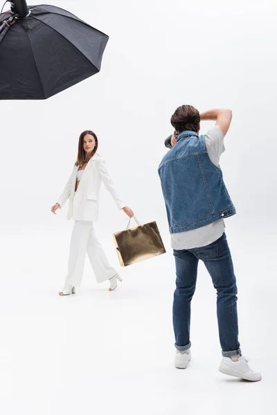 Back view of photographer taking photo of model in white suit walking with golden shopping bag on white — Stock Photo