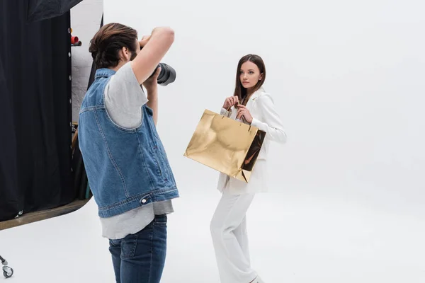 Jeune modèle en costume blanc posant avec sac à provisions doré près du photographe sur blanc — Photo de stock