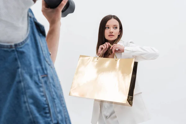 Fotógrafo desfocado tirando foto do modelo moderno com saco de compras dourado isolado em branco — Fotografia de Stock