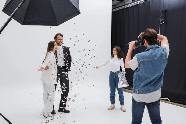 Smiling models in suits posing near photographer and assistant throwing confetti — Stockfoto