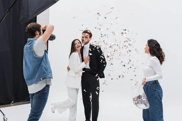 Modelos alegres en trajes posando durante la sesión de fotos cerca de asistente lanzando confeti - foto de stock