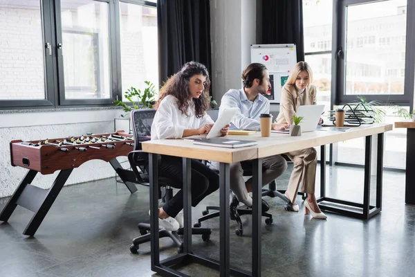 Junge Geschäftsleute sitzen mit Geräten am Schreibtisch, während sie gemeinsam im Büro arbeiten — Stockfoto