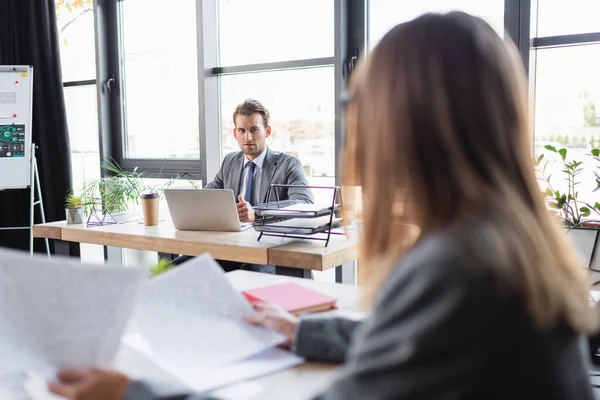 Businessman looking at blurred colleague while sitting at desk near laptop — Stock Photo