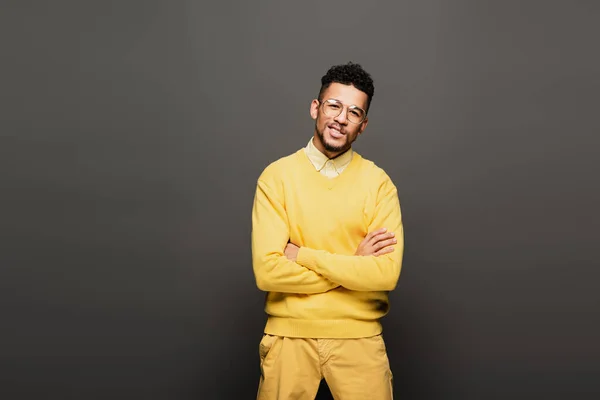 Homme afro-américain joyeux en tenue jaune et lunettes debout avec les bras croisés sur gris foncé — Photo de stock