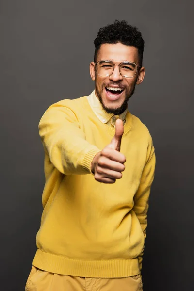 Excited african american man in yellow outfit and glasses showing thumb up isolated on grey — Stock Photo