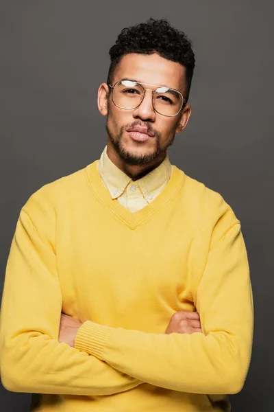 Hombre afroamericano serio en traje amarillo y gafas de pie con brazos cruzados aislados en gris - foto de stock