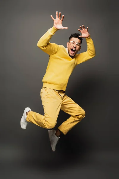 Asombrado hombre afroamericano en gafas y traje amarillo gestos mientras salta sobre gris oscuro - foto de stock