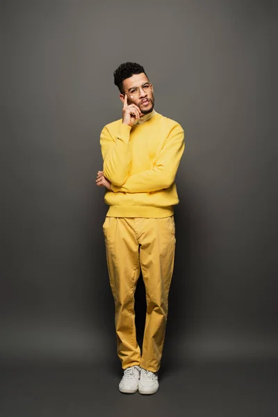 Homem americano africano pensativo em óculos e roupa amarela olhando para a câmera no cinza escuro — Fotografia de Stock