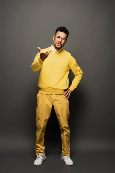 Displeased african american man in glasses and yellow outfit gesturing on dark grey — Stock Photo