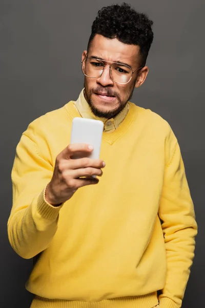 Worried african american man in glasses looking at smartphone isolated on grey — Stock Photo