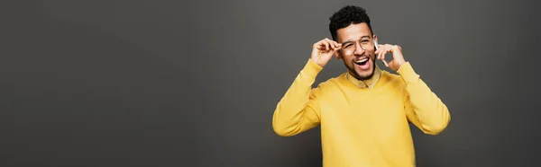 Hombre afroamericano feliz ajustando gafas y hablando en el teléfono inteligente en gris oscuro, bandera - foto de stock