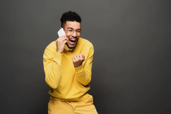 Excited african american man adjusting glasses and talking on smartphone on dark grey — Stock Photo