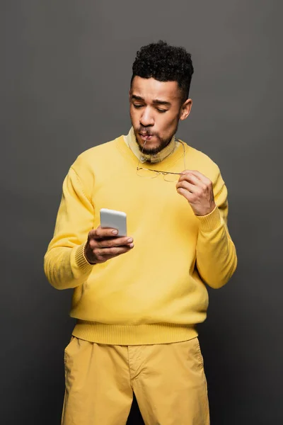 African american man holding glasses near lips and using smartphone isolated on grey — Stock Photo