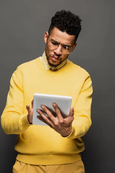 Giovane uomo afroamericano in occhiali utilizzando tablet digitale offuscata isolato su grigio — Foto stock