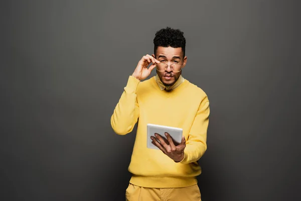 Focused african american man adjusting glasses and holding digital tablet on dark grey — Stock Photo