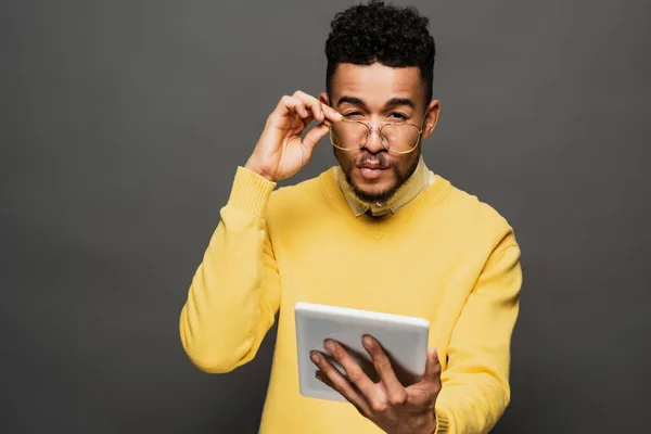 Hombre afroamericano enfocado ajustando gafas y sosteniendo la tableta digital en gris oscuro - foto de stock