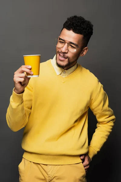 Cheerful african american man in glasses looking at paper cup on dark grey — Stock Photo