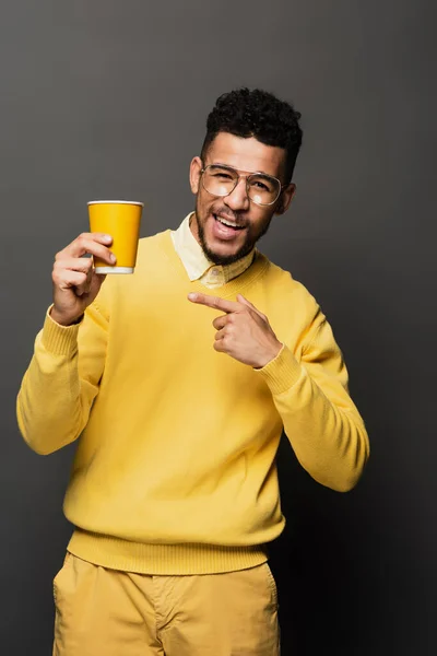 Hombre afroamericano excitado en gafas apuntando con el dedo a la taza de papel en gris oscuro - foto de stock