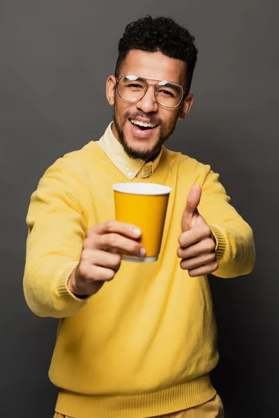 Felice uomo afroamericano in bicchieri tenendo tazza di carta e mostrando pollice in alto isolato sul grigio — Foto stock