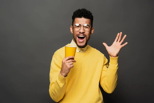 Hombre afroamericano excitado en gafas sosteniendo una taza de papel y haciendo gestos en gris oscuro - foto de stock