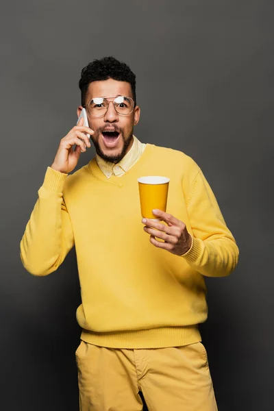 Homme afro-américain en lunettes et pull jaune tenant tasse en papier et parlant sur smartphone sur gris foncé — Photo de stock