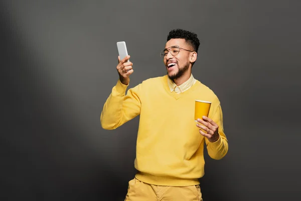 Heureux homme afro-américain en lunettes et pull jaune tenant tasse en papier et regardant smartphone sur gris foncé — Photo de stock