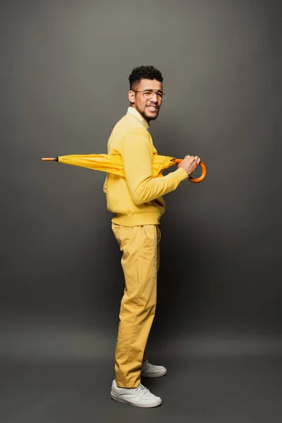 Homme afro-américain mécontent en lunettes et tenue jaune tenant parapluie sur gris foncé — Photo de stock
