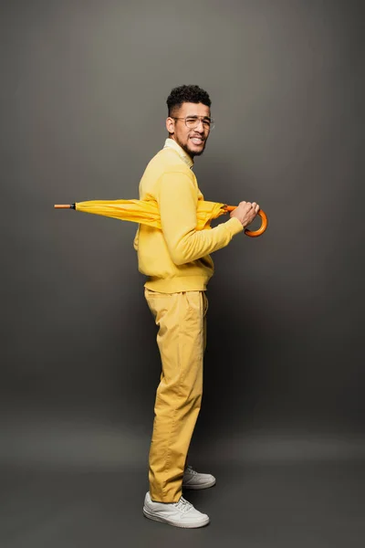 Displeased african american man in glasses and yellow outfit holding umbrella on dark grey — Stock Photo