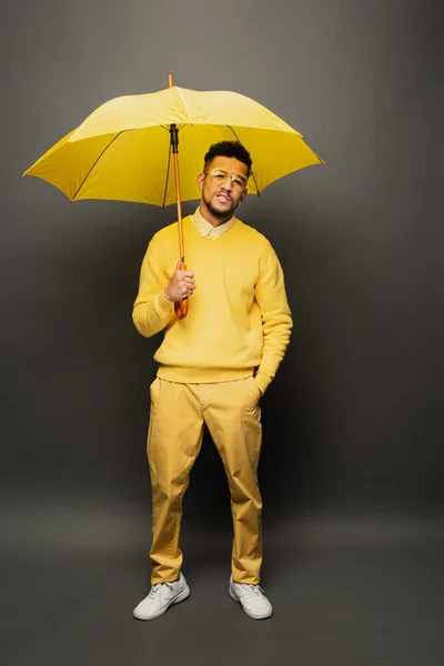 Curly african american man in glasses and yellow outfit standing with hand in pocket under umbrella on dark grey — Stock Photo