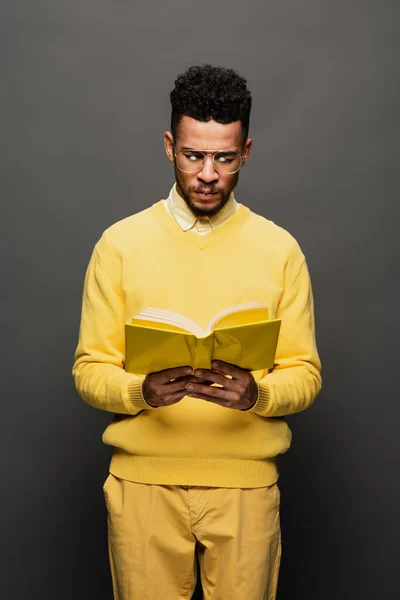 African american man in glasses and yellow outfit reading book on dark grey — Stock Photo