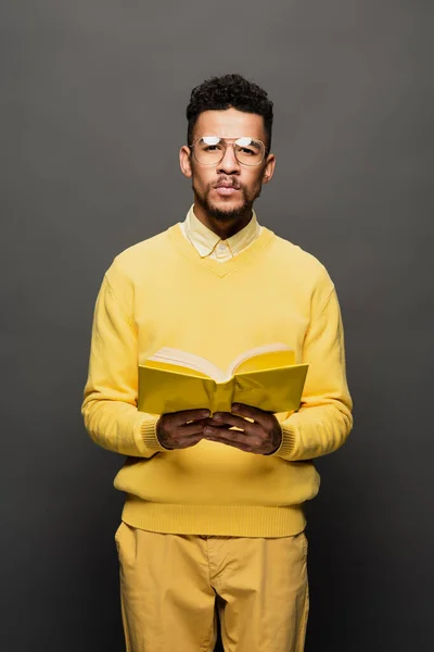 African american man in glasses and yellow outfit holding book on dark grey — Stock Photo