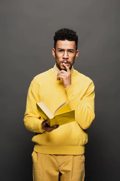 Pensive african american man in glasses and yellow outfit holding book on dark grey — Stock Photo