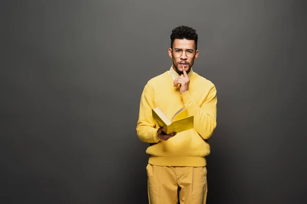 Homme afro-américain réfléchi dans des lunettes et tenue jaune tenant livre sur gris foncé — Photo de stock