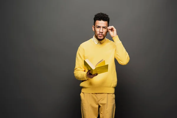 Focused african american man adjusting glasses and holding book on dark grey — Stock Photo