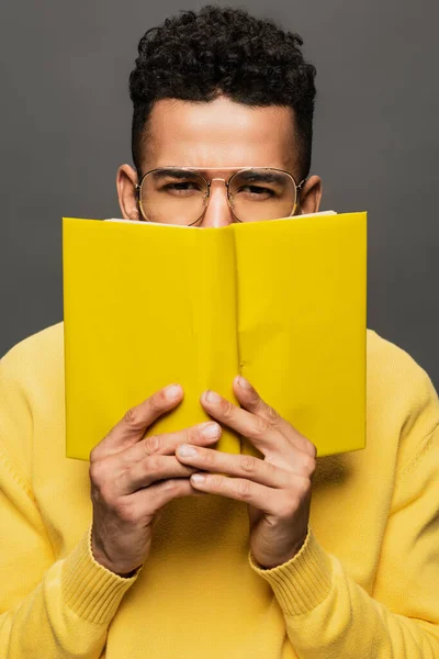 African american man in glasses and yellow outfit holding book isolated on grey — Stock Photo