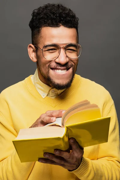 Alegre afroamericano hombre en gafas y suéter amarillo celebración libro aislado en gris oscuro - foto de stock