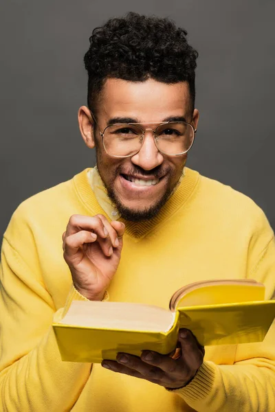 Sly african american man in glasses and yellow sweater holding book and smiling isolated on grey — Stock Photo