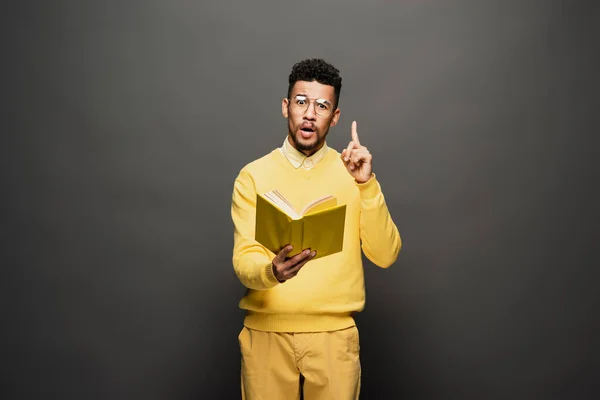 Surprised african american man in glasses and yellow outfit holding book while showing idea sign on dark grey — Stock Photo