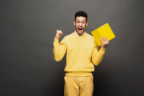 Excited african american man in glasses and yellow outfit holding book on dark grey — Stock Photo