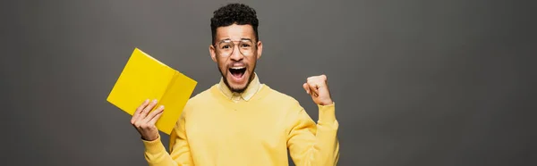 Excited african american man in glasses and yellow outfit holding book on dark grey, banner — Stock Photo