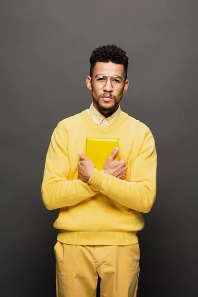 Curly african american man in glasses and yellow outfit holding book on dark grey — Stock Photo