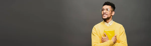 Sonriente hombre afroamericano en gafas y traje amarillo sosteniendo libro en gris oscuro, bandera - foto de stock