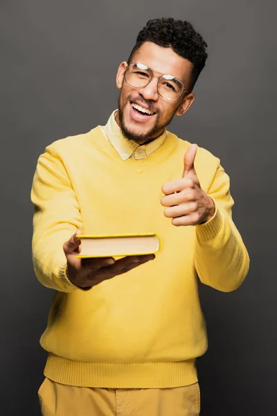 Heureux homme afro-américain en lunettes et pull jaune montrant pouce vers le haut et tenant livre isolé sur gris — Photo de stock